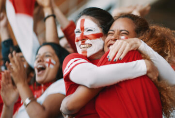 Women embracing in stadium crowd wearing England kit and face paint in celebration