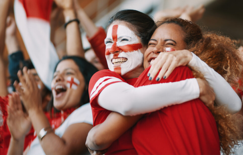 Women embracing in stadium crowd wearing England kit and face paint in celebration