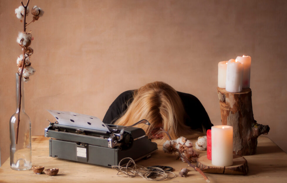 woman rests head on table in defeat with typewriter in front of her on the desk