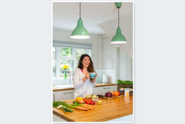 Woman standing at kitchen counter with cup of tea, laughing with vegetables laid out in front