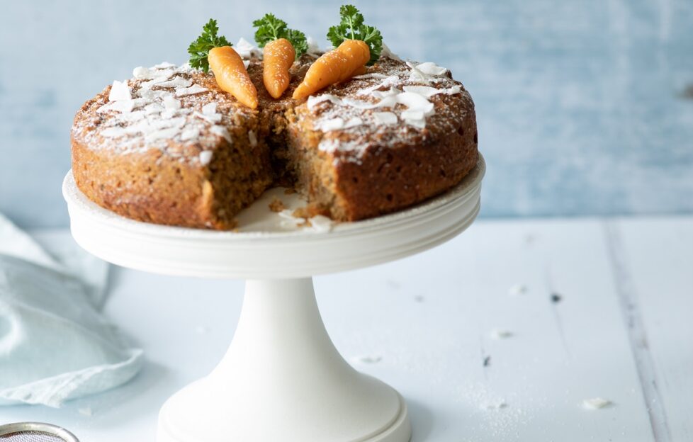 Carrot, almond and coconut cake on a white cake stand with decorative carrots on top and a slice taken out