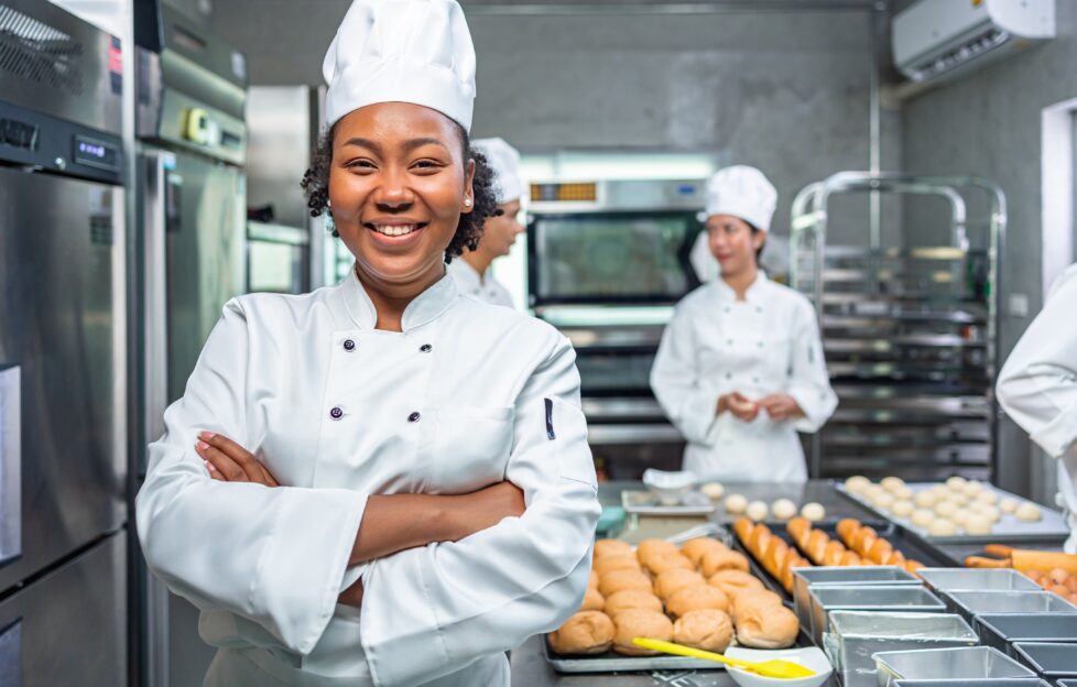 Smiling lady chef in hotel kitchen with other chefs and recipe ingredients.