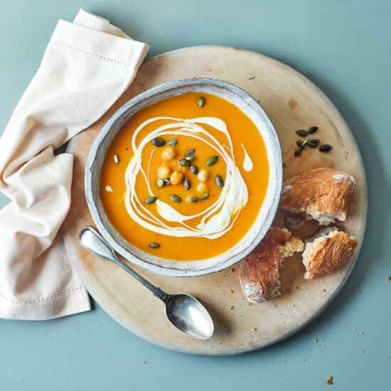 Top down flatlay view of pumpkin soup in a bowl with drizzled cream and sat on a plate with crusty bread to serve