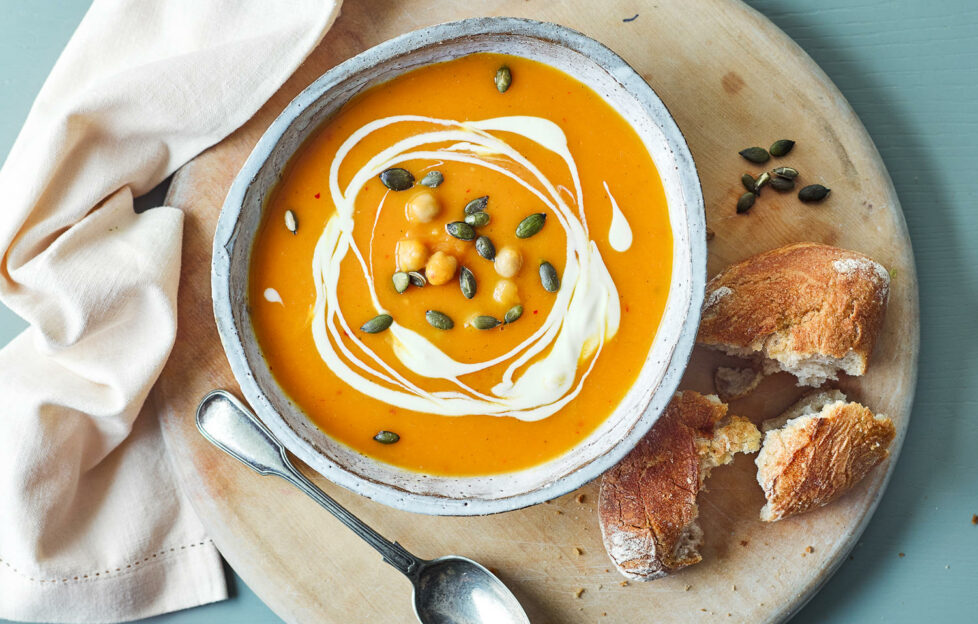 Top down flatlay view of pumpkin soup in a bowl with drizzled cream and sat on a plate with crusty bread to serve