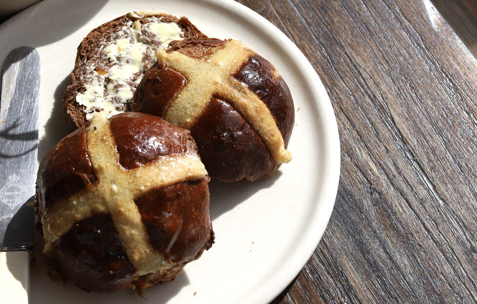A plate of hot cross buns on a table in the sunshine, one halved and spread with butter