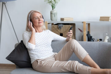 Grey haired woman reclining on sofa listening to music through headphones