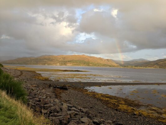 A view of Lochcarron from the main street out across the loch into the hills as the golden sun sets, with a rainbow peeking at the right side of the frame