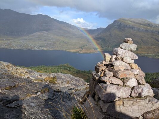 Up the trail of Beinn Eighe Nature Reserve, with a view of Loch Maree, a rainbow bursting from behind a stone waymark