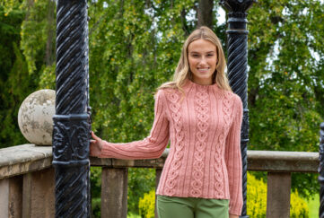 Blonde model wearing salmon pink cable knit sweater and green trousers with a backdrop of trees
