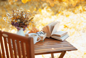 A wood table with bouquet of flowers, cup of tea, books and reading glasses against a backdrop of autumn leaves