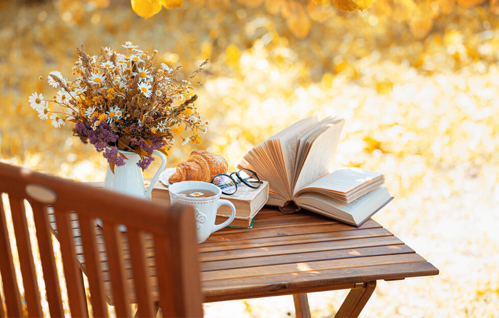 A wood table with bouquet of flowers, cup of tea, books and reading glasses against a backdrop of autumn leaves