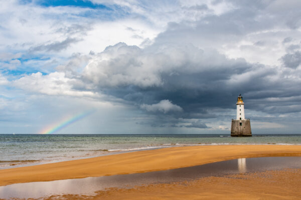 A dramatic view from Fraserburgh beach out to sea with the lighthouse, thick clouds and a rainbow on the horizon