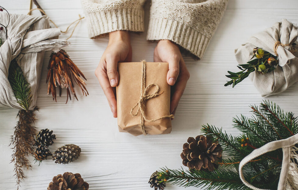 Flatlay of hands holding recyclable wrapped gift and sustainable Christmas items, pinecones and pine surrounding