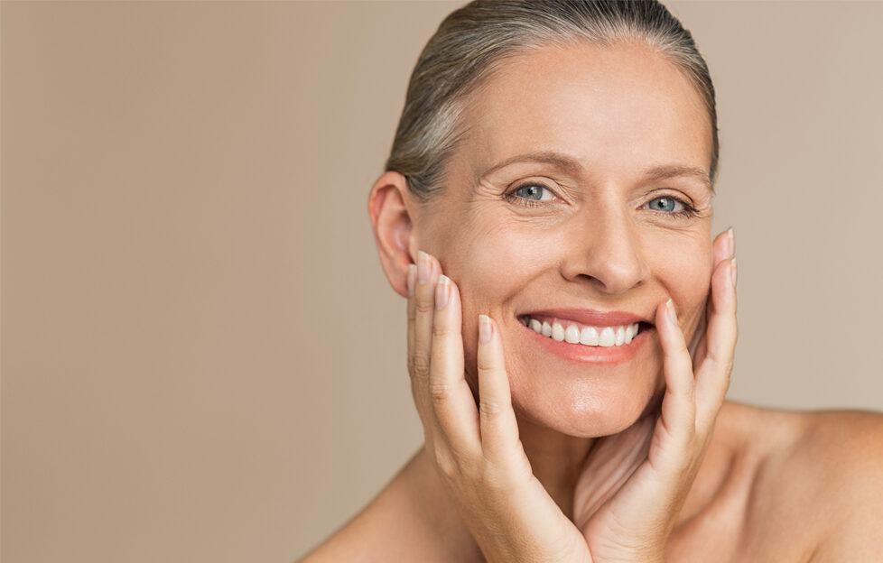 Mature woman smiling and cupping her face with her hands, grey hair pulled back in a ponytail