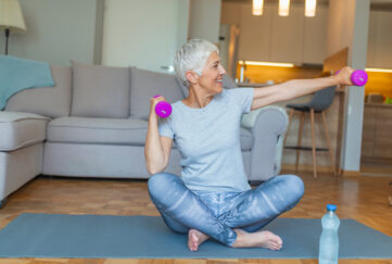 Grey haired woman sitting on a yoga mat in active clothes, smiling as she stretches with dumbells