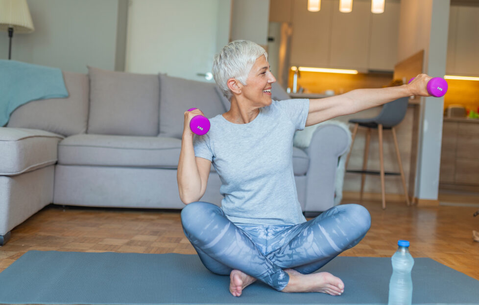 Grey haired woman sitting on a yoga mat in active clothes, smiling as she stretches with dumbells