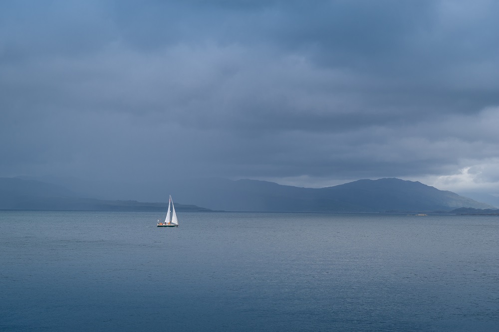 A lone sailing boat on a gloomy grey sea with misty hills in the background