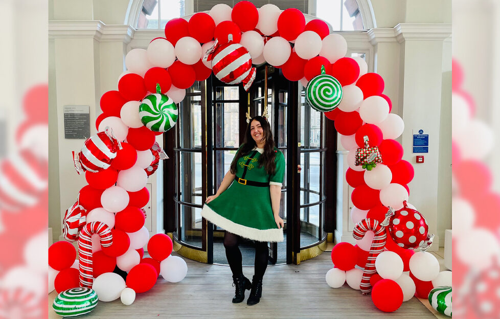 Jacqueline in a green elf dress standing underneath a Christmas balloon arch