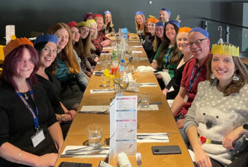 The People's Friend team sat up a long table in a restaurant having Christmas dinner with party hats and crackers, smiling at the camera