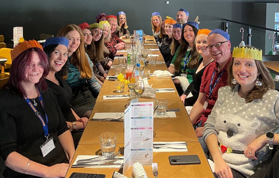 The People's Friend team sat up a long table in a restaurant having Christmas dinner with party hats and crackers, smiling at the camera