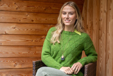 Blonde woman sitting in front of wood panel walls wearing green diamond knitted sweater with green shirt collar underneath