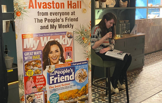 Kirsty reading a story at the Warner Alvaston Hall holiday next to a banner of My Weekly and The People's Friend