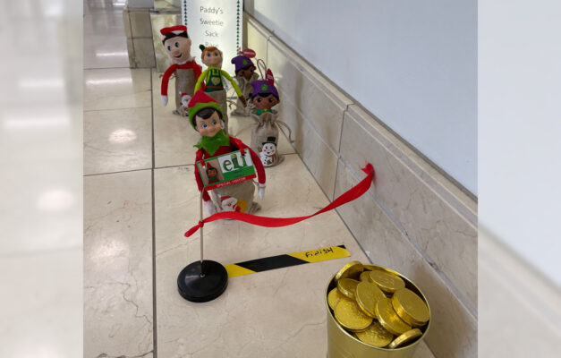 Paddy the Elf doll and other toys behind in a line wearing sacks in a 'sack race' to a red finish line and a pot of gold at the end on the office floor