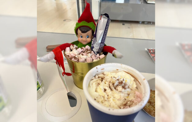 Paddy the Elf doll sitting in a tub of marshmallows and chocolate next to a hot chocolate from the office