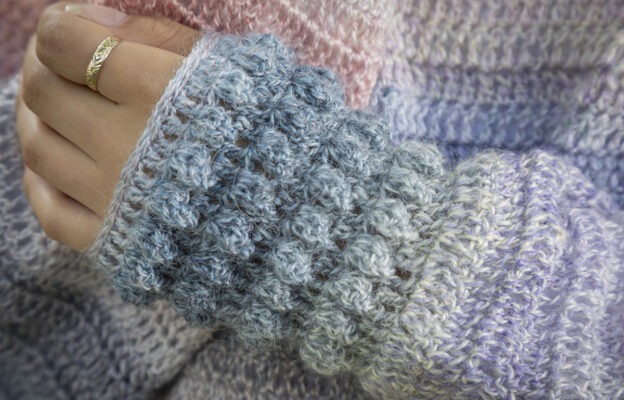 Close up of multi pastel coloured crochet popcorn pattern sweater sleeve cuff worn by model with gold ring on first finger