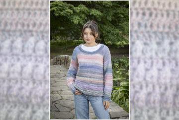 Brown haired model standing in front of bridge and grassy bank with pastel coloured popcorn pattern crochet sweater and light blue jeans