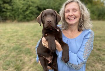 Jackie (with shoulder length blonde hair wearing a blue top smiling) holding Logan a chocolate labrador in her arms looking directly at the camera in a grassy field