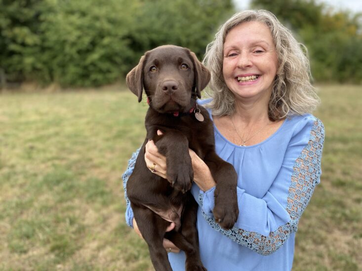 Jackie (with shoulder length blonde hair wearing a blue top smiling) holding Logan a chocolate labrador in her arms looking directly at the camera in a grassy field