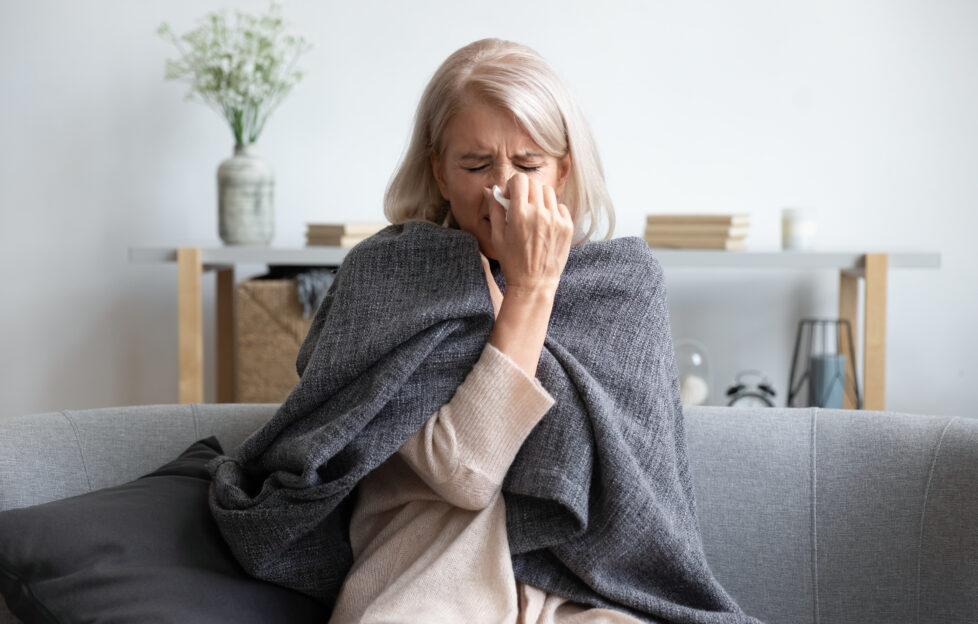 Woman with blonde/white hair huddled in a blanket sneezing into tissue