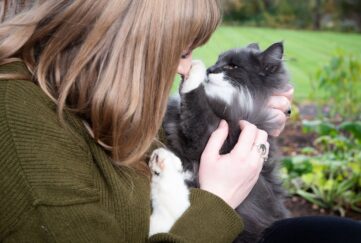 Blonde woman wearing green coat sat in the grass holding black and white fluffy cat who's pressing their paw against her nose