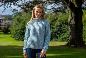 Blonde woman wearing knitted cosy ribbed sweater in light shade of blue and dark blue jeans, standing in front of the shade of a tree and bushes