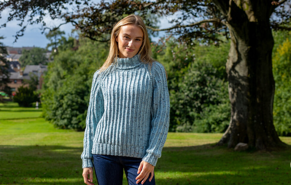 Blonde woman wearing knitted cosy ribbed sweater in light shade of blue and dark blue jeans, standing in front of the shade of a tree and bushes