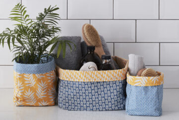 Three grey and mustard patterned fabric storage boxes of different sizes, one holidng a plant, the other holding eco cleaning supplies, in front of a white tiled wall