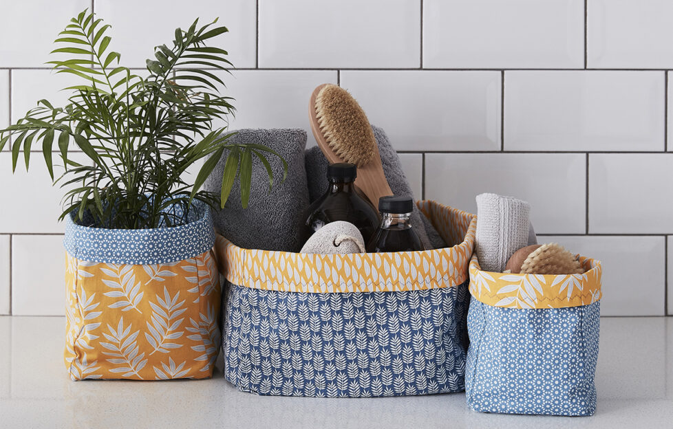 Three grey and mustard patterned fabric storage boxes of different sizes, one holidng a plant, the other holding eco cleaning supplies, in front of a white tiled wall