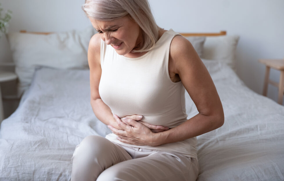 Mature silver haired woman sat on the bed holding stomach in clear discomfort and feeling sick