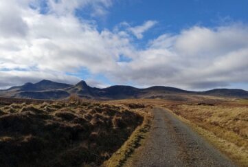 A road in Skye with hills in the background and a blue sunny sky with clouds.