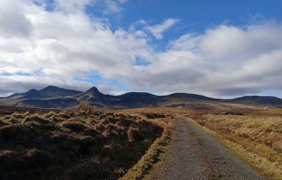 A road in Skye with hills in the background and a blue sunny sky with clouds.