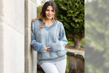 Brown haired model leaning against a wall wearing cosy light blue knitted v-neck sweater and white jeans