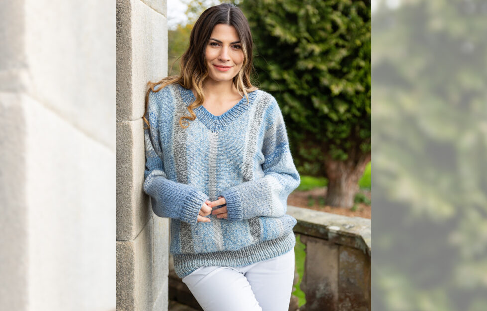 Brown haired model leaning against a wall wearing cosy light blue knitted v-neck sweater and white jeans