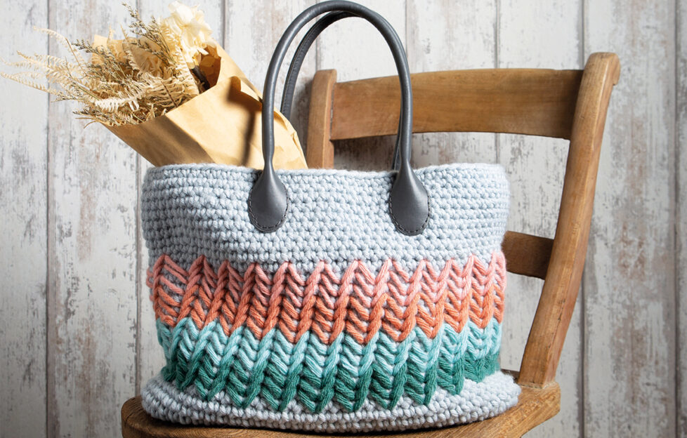 Grey crochet handbag witith pink, orange, blue and green zig-zagged stripes across and black handles on top of a wooden chair with dried flower bouquet inside