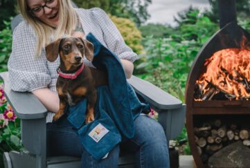 Blonde woman sitting by a fire pit laughing with a brown small dog on her lap cleaning them with a towel