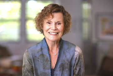 A still shot of Judy Blume in the documentary 'Judy Blume Forever'. Image features Judy Blume against a blurred home background. She is wearing a dusty grey and blue sweater and smiling for the camera.