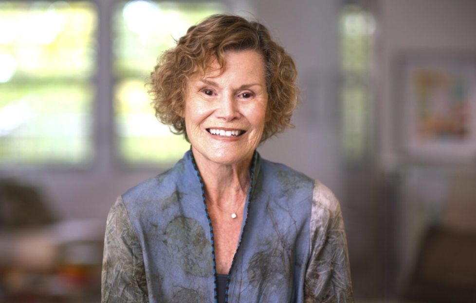 A still shot of Judy Blume in the documentary 'Judy Blume Forever'. Image features Judy Blume against a blurred home background. She is wearing a dusty grey and blue sweater and smiling for the camera.
