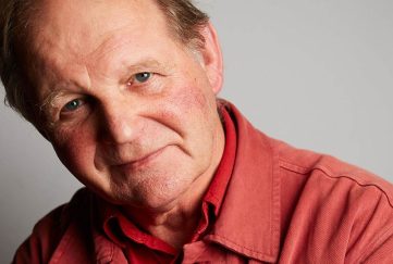 Image shows former Children's Laureate and current BookTrust Michael Morpurgo. He is softly smiling and wearing a light, red shirt.