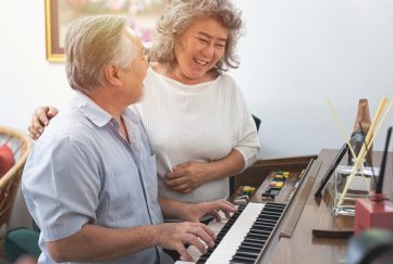 Playing a musical instrument is good for our brain health. Image feature an elderly Asian couple. The husband is playing the piano while loving looking at his wife who stands next to him, hugging his shoulders.