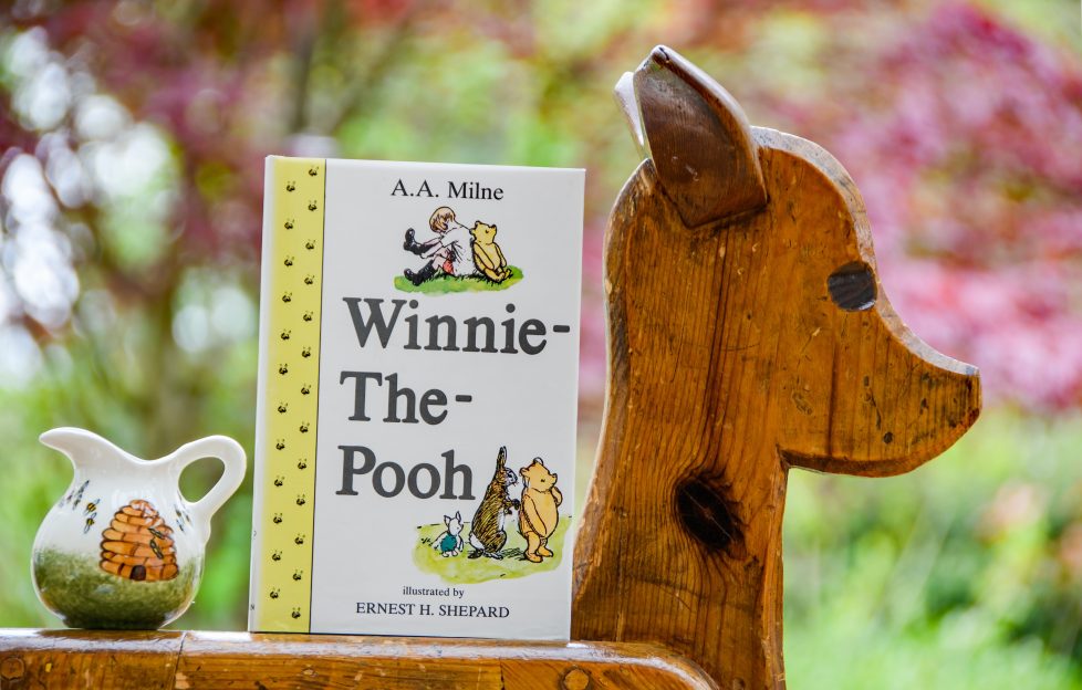 Winnie The Pooh quotes. Image features: Winnie The Pooh hardback book propped up on a ornamental wooden deer carving. A small jar of honey with a lovely bee illustration sits next to it.